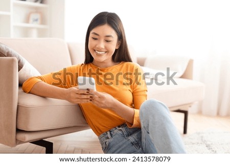 Cute pretty young asian woman sitting on floor next to couch at home, using phone and smiling, texting her friends or lover, enjoying newest mobile app, scrolling, shopping online, copy space