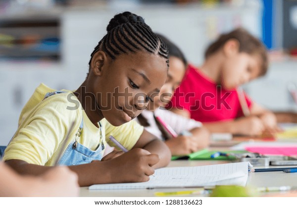 Cute pupil writing at desk in classroom at the\
elementary school. Student girl doing test in primary school.\
Children writing notes in classroom. African schoolgirl writing\
during the lesson.