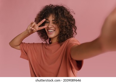 cute radiant lady makes selfie showing two fingers near her face on pink background. dark-haired, curly, smiling African woman with snow-white smile blinks one eye.