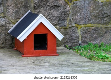 Cute Red Doghouse With White Roof Trim On A Weathered Wood Deck Against A Stone Wall
