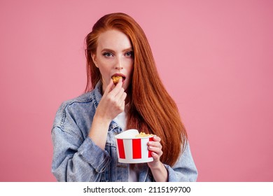 Cute Red Long Foxy Haired Ginger Woman Holding Packaging Cardboard Box Bucket With French Fries And Eating Chicken Crunchy Nuggets In Pink Studio