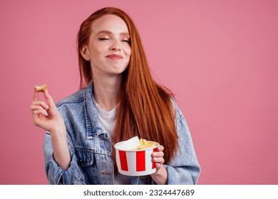 cute red long foxy haired ginger woman holding packaging cardboard box bucket with french fries and eating chicken crunchy nuggets in pink studio