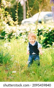 Cute red-haired baby boy is standing on a green grass in the park on a sunny day