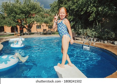 Cute Red-haired Girl Diving In Pool From Springboard. Child Kid Enjoying Having Fun In Swimming Pool On Home Backyard. Summer Outdoor Water Activity For Kids. 