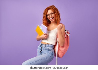 Cute redhead curly girl student teenager with a backpack holds notepads and notebooks and shows the gesture yes as a winner isolated on a purple background.
