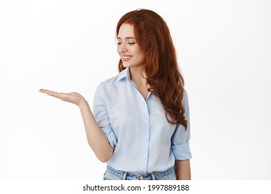 Cute redhead woman looking at open palm and smiling at product, holding item copy space in hand, standing in office blouse against white background - Powered by Shutterstock