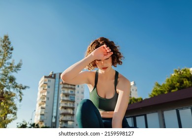 Cute Redhead Woman Sitting On Gras Wearing Sports Bra And Blue Yoga Pants After Jogging Catching Her Breath, Wiping Her Sweat From Her Forehead.