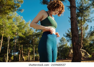 Cute redhead woman wearing green sports bra and blue yoga pants standing on city park, outdoors she holds hands at the waist. Flat stomach, healthy life and outdoor sport concepts.