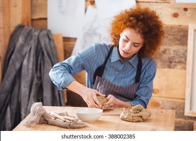 Cute redhead young female ceramist working with clay and making dishes by hands in pottery studio