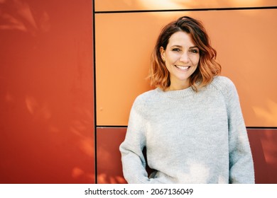 Cute relaxed young woman with a genuine beaming smile and happy friendly expression standing against a colorful orange exterior wall with copyspace - Shutterstock ID 2067136049
