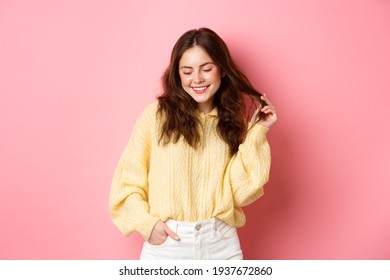 Cute and romantic brunette woman playing with her hair, blushing and looking down with coquettish smile, standing flirty against pink background - Shutterstock ID 1937672860