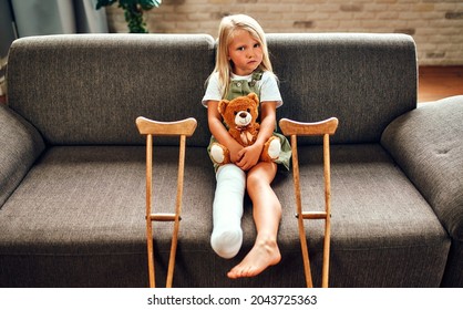A cute sad little girl with a broken leg in a cast, sits on the couch hugging a teddy bear at home. There are crutches near the sofa for quick rehabilitation.