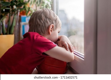 Cute Sad Little Kid With Football Ball Sitting On The Window Sill And Looking On The Street, Dream About Outdoor Activity During Quarantine Lockdown, Indoor Lifestyle, Concept Of Sorrow