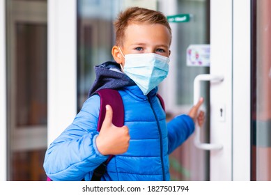 Cute School Boy 9s Old Wears Casual Clothes And Backpack Face Mask Near School. Kid Shows Thumbs Up And Holds Or Opens The Door Handle