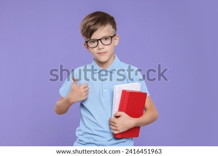 Cute schoolboy in glasses holding books and showing thumbs up on violet background