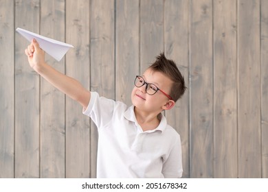 Cute Schoolboy Making And Launching A Paper Airplane Wearing Glasses And A White T-shirt On Wooden Background