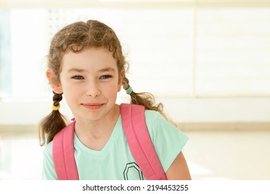 Cute Schoolgirl With A Backpack Stands In School Hall. 7 Year Old Caucasian Girl Looking Away. Back To School, Education, Learning Concept. Indoor. Copy Space.