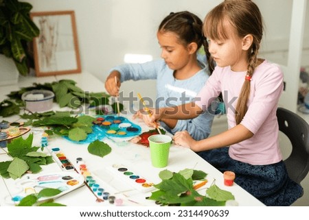 Cute schoolgirl with scissors cutting dry oak leaf while helping her teacher with decorations for holiday at lesson
