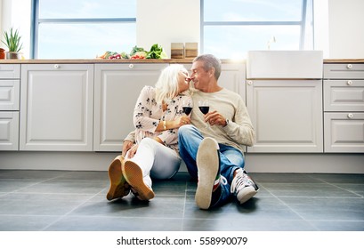 Cute senior husband and wife kissing each other while seated on floor in kitchen with bright windows and large cabinets - Powered by Shutterstock