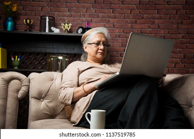 Cute Senior Indian/asian Lady Sitting On Recliners Chair Or Sofa Reading Book Or Using Tab Or Laptop Computer