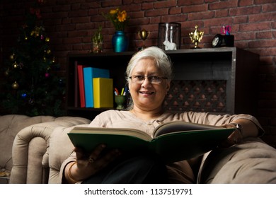 Cute Senior Indian/asian Lady Sitting On Recliners Chair Or Sofa Reading Book Or Using Tab Or Laptop Computer