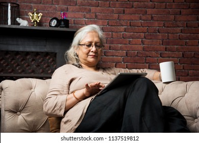 Cute Senior Indian/asian Lady Sitting On Recliners Chair Or Sofa Reading Book Or Using Tab Or Laptop Computer