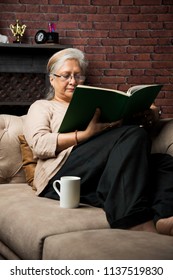 Cute Senior Indian/asian Lady Sitting On Recliners Chair Or Sofa Reading Book Or Using Tab Or Laptop Computer