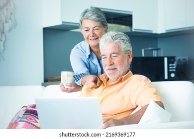 Cute Senior Man And Woman Using Computer At Home. Man Sitting On The Sofa And Woman Behind Him Watching The Laptop Display. Mature People Surfing The Web Indoor. Elderly Real Lifestyle