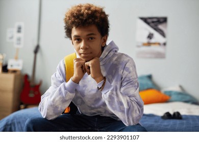 Cute serene teenage boy in casualwear keeping hands by chin while sitting on double bed in his bedroom at leisure and looking at camera