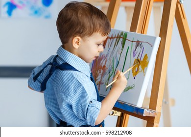 Cute, serious and focused, three years old boy in blue shirt and jeans apron drawing on canvas standing on the easel. Concept of early childhood education, painting, talent, happy family or parenting - Powered by Shutterstock