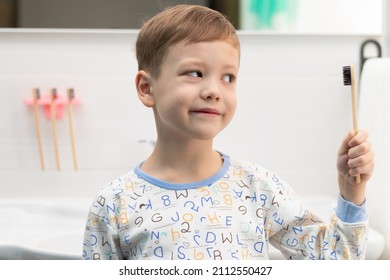 A cute seven-year-old boy in pajamas with a bamboo toothbrush in his hand before going to bed at home in the bathroom against the backdrop of a washbasin with a faucet. Selective focus. Portrait