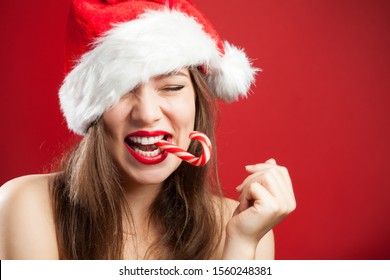 Cute sexy caucasian brunette young woman dressed as Mrs. Santa Claus, smiling with joy, biting a candy cane with eyes closed, Christmas or New Years Eve 2022 concept, copy space on red backdrop 