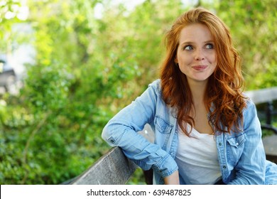 Cute Sexy Young Redhead Woman With A Quirky Smile Sitting Relaxing On A Wooden Bench In A Park Glancing To The Side