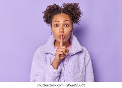 Cute shocked young woman makes silence gesture presses index finger over lips shows hush sign says shh dressed in outerwear spreads gossips isolated over purple background. Body language concept