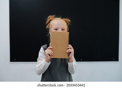 Cute Shy Schoolgirl Peeking Over Textbook, Holding In Over Her Face. Standing In Front Of A Balckboard In A Classroom.