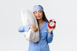 Cute And Silly Asian Girl In Blue Pyjamas And Sleeping Mask, Sleeping On Pillow With Closed Eyes And Showing Alarm Clock, Having Sweet Dreams As Forgot Set Up Alarm, White Background