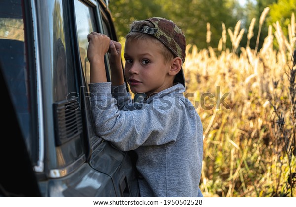 a cute six year old boy stands by the car\
against the background of autumn grass. film grain effect.\
horizontal orientation.