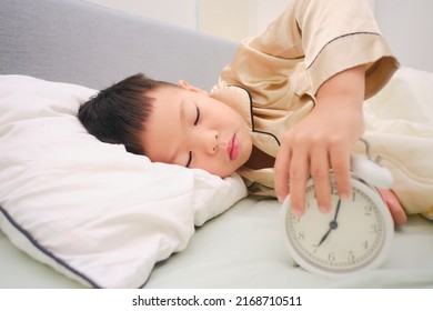 Cute Sleepy Little Asian Kindergarten Boy Child Lying On The Bed And Stopping, Turn Off Alarm Clock In The Bedroom At Home, Kid Hates Waking Up Early In The Morning, Selective Focus At The Boy