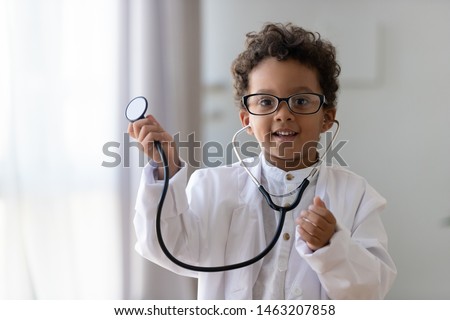 Cute small african american kid boy wear medical uniform glasses holding stethoscope playing doctor, happy funny little mixed race preschool child pretending pediatrician looking at camera, portrait