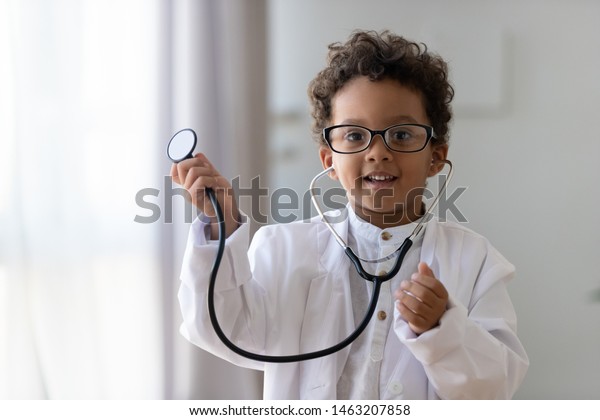 Cute small african american kid boy wear medical\
uniform glasses holding stethoscope playing doctor, happy funny\
little mixed race preschool child pretending pediatrician looking\
at camera, portrait