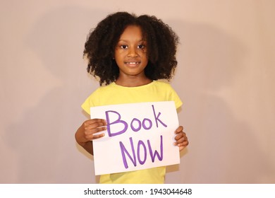 Cute Small Black Child Holding Sign With Words Book Now Indoors Plain Grey Background