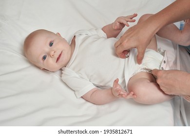 Cute Small Boy Lying At Bed. Childhood Concept. Light Background. Serious Child. Copyspace. Stay Home. Onesie Mockup. Mother Hands Close Buttons. Look At Camera. White Clothes