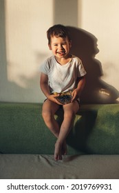 Cute Small Boy Is Smiling At Camera While Eating Blueberries Sitting On The Couch At Home