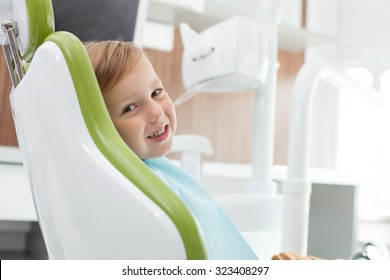 Cute small boy is visiting dentals doctor. He wants to treat his teeth. The kid is sitting in medical chair with joy. He is looking at camera and smiling