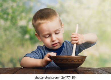 Cute small child boy eats his food from clay bowl with wooden spoon outdoors