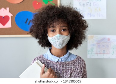 Cute Small Elementary Reopen School Pupil African American Kid Child Girl Wearing Face Mask Looking At Camera Standing In Classroom. Children Safety For Covid Protection, Headshot Close Up Portrait.