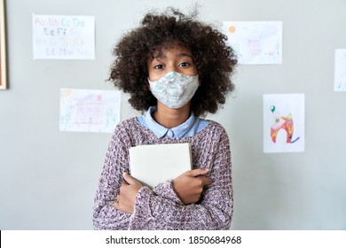 Cute Small Elementary Reopen School Pupil African American Kid Child Girl Wearing Face Mask Holding Book Looking At Camera Standing In Classroom. Children Safety Covid Prevention Protection Concept.