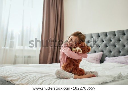 A cute small girl smiling embracing a teddybear while sitting barefoot on a huge bed wearing pyjamas in a bright spacy bedroom at home