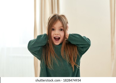 A cute small girl surprised with her eyes and mouth wide open and hands covering her ears looking into a camera in a big room at home alone. Children 