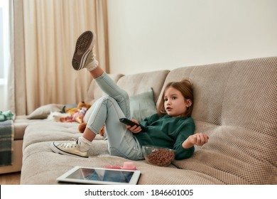 A Cute Small Girl Wondering What To Watch On A TV Holding A TV Remote Sitting On A Sofa In A Big Room With A Bowl Of Cereal Balls Being Alone At Home. Childrens Leisure Activities